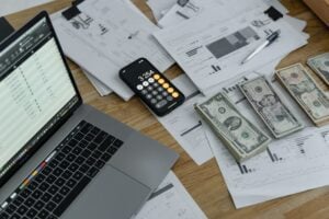 A laptop sits on a desk next to budgeting paperwork, a calculator and stacks of paper money