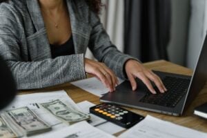 A person works on a laptop next to paperwork, a calculator and a stack of money