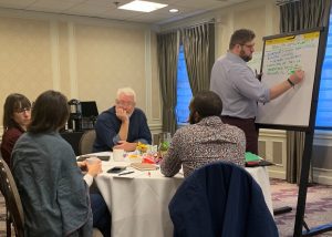 Colin Groth writing on a board while working with a group of other people who are seated at a table