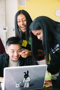 A teenage boy and two teenage girls gather around a laptop, smiling and laughing.