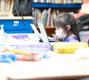 A young girl with light-colored skin and brown hair sits at a table, learning how to use a laptop. She is wearing a white face mask. 