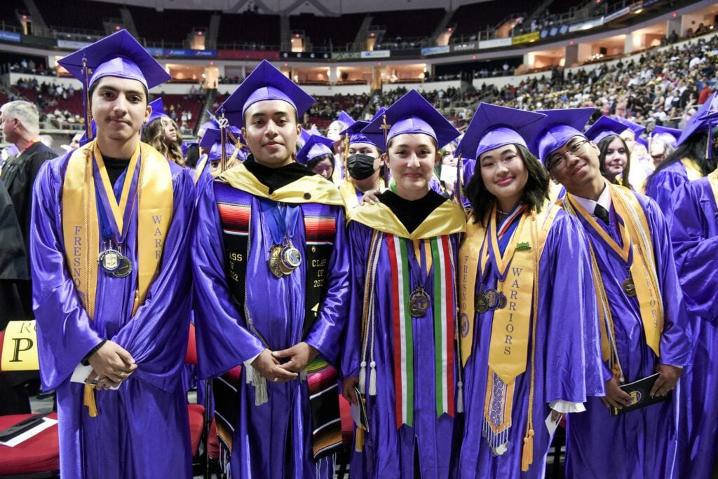 A group of five Fresno High School graduates smile at the camera. They're wearing purple graduation caps and gowns, as well as assorted medals, stoles and cords. 