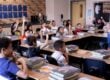 17 elementary school students sit at tables in a classroom. Most of them have their hands raised, waiting to be called on.