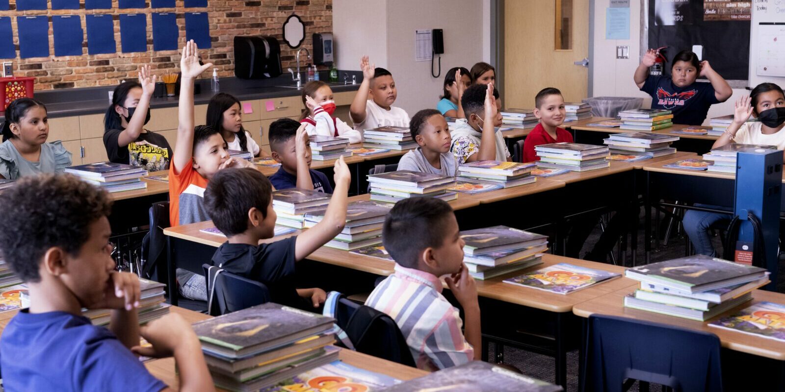 17 elementary school students sit at tables in a classroom. Most of them have their hands raised, waiting to be called on.