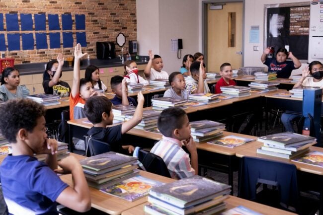 17 elementary school students sit at tables in a classroom. Most of them have their hands raised, waiting to be called on.