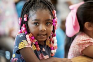 A pre-k girl smiles shyly at the camera. She is Black and has braided hair with red, pink and white beads at the ends.