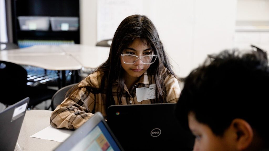 A young girl wearing glasses looks down at a laptop.