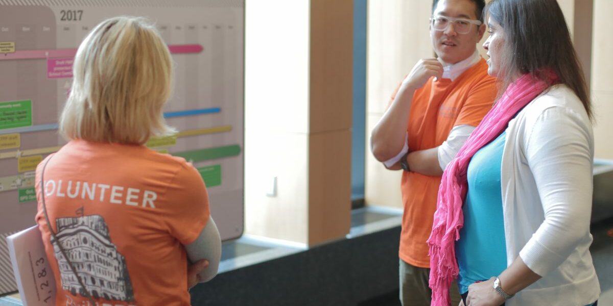 A group of three people stand discussing a timeline that's posted on the wall