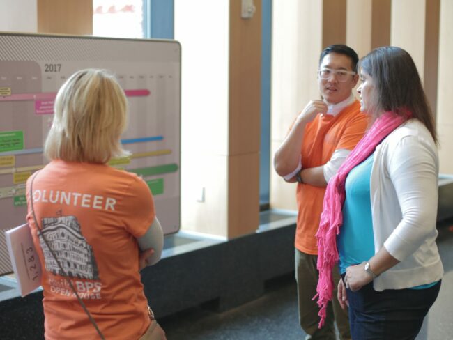 A group of three people stand discussing a timeline that's posted on the wall