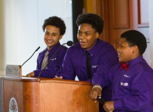 Three black boys wearing purple shirts stand behind a podium. One of them is speaking into a microphone.