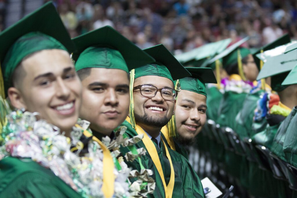 Four high school graduates sit in a row, smiling at the camera. They're wearing green caps and gowns.