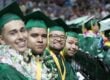 Four high school graduates sit in a row, smiling at the camera. They're wearing green caps and gowns.