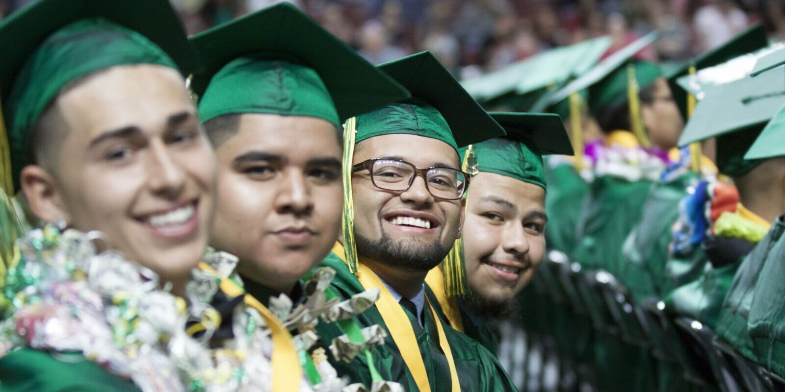 Four high school graduates sit in a row, smiling at the camera. They're wearing green caps and gowns.