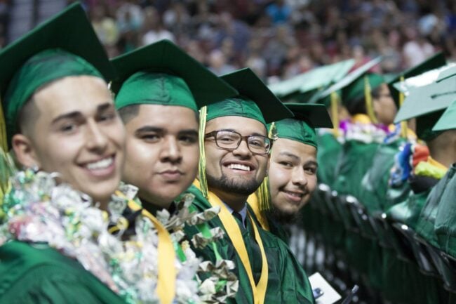 Four high school graduates sit in a row, smiling at the camera. They're wearing green caps and gowns.