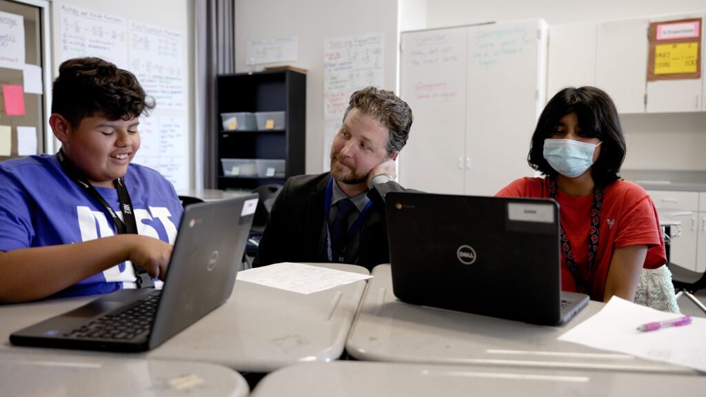 A male teacher sits between two students who are working on laptops