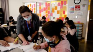 A teacher wearing a blue face mask leans over a table to help a female student who is also wearing a blue face mask.