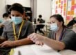 A female teacher wearing a blue medical face mask kneels next to a table, helping a male student who is also wearing a blue medical face mask.