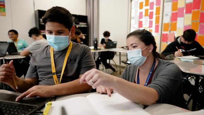 A female teacher wearing a blue medical face mask kneels next to a table, helping a male student who is also wearing a blue medical face mask.