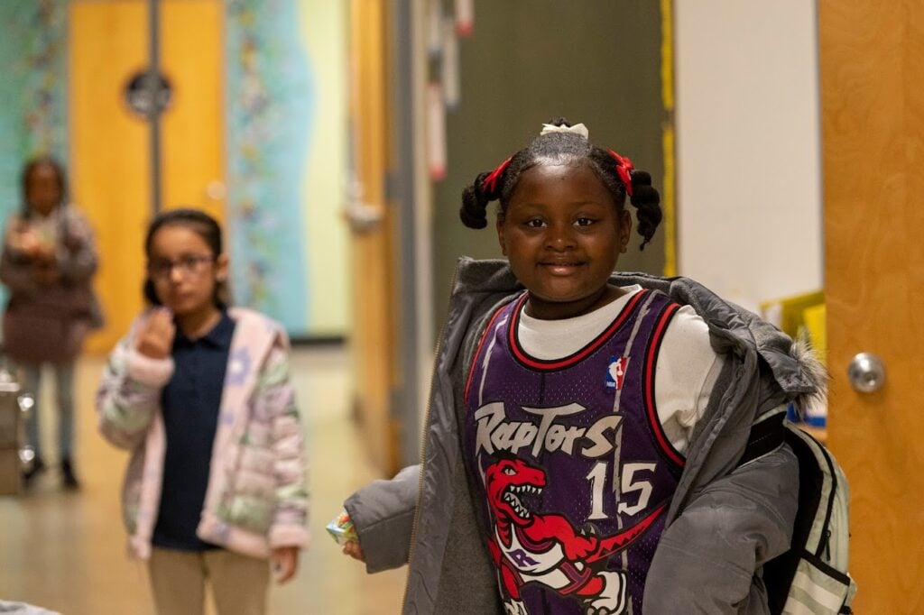 A young girl stands in the hallway, smiling at the camera. She's wearing a Toronto Raptors jersey.