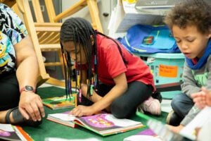 A young girl kneels on the ground reading a book. She has long braids with different colored beads on the bottom.