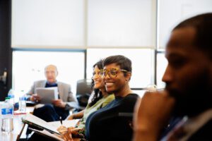 In the focus of the photo, two women sit next to each other at a table, smiling. In the background and foreground, two other people sit at the table, out of focus. 