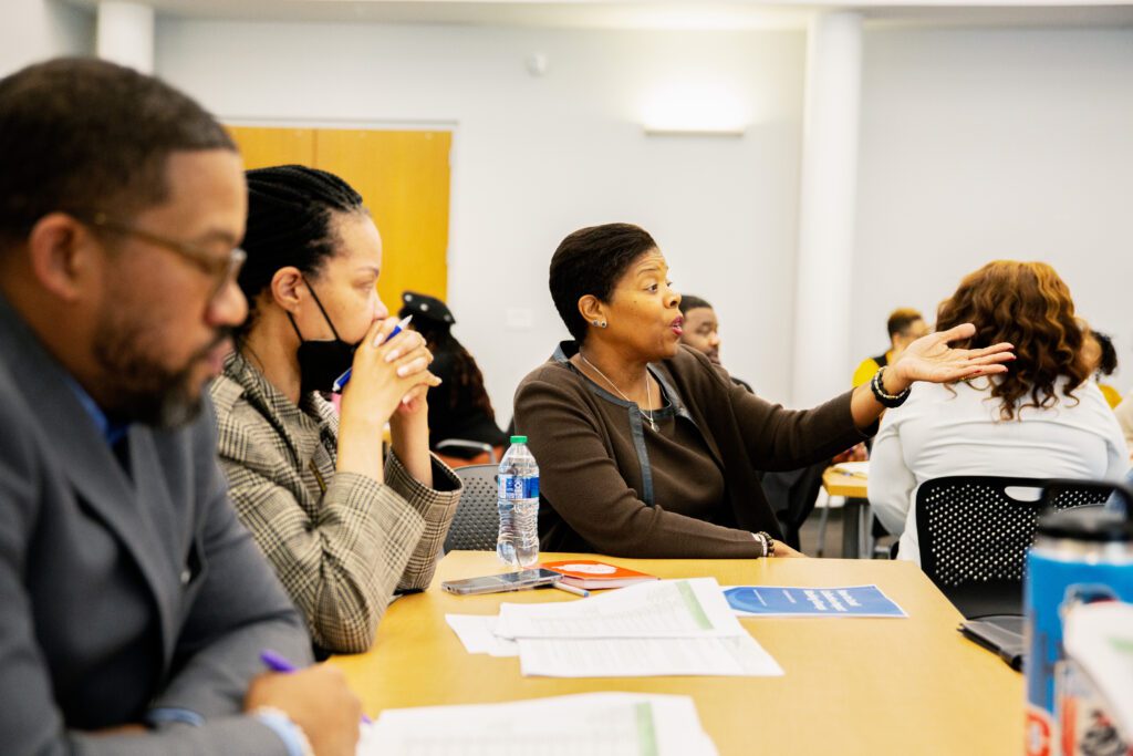 A woman sits at the head of a table, speaking with her arm outstretched. Two others sit next to her, looking forward.