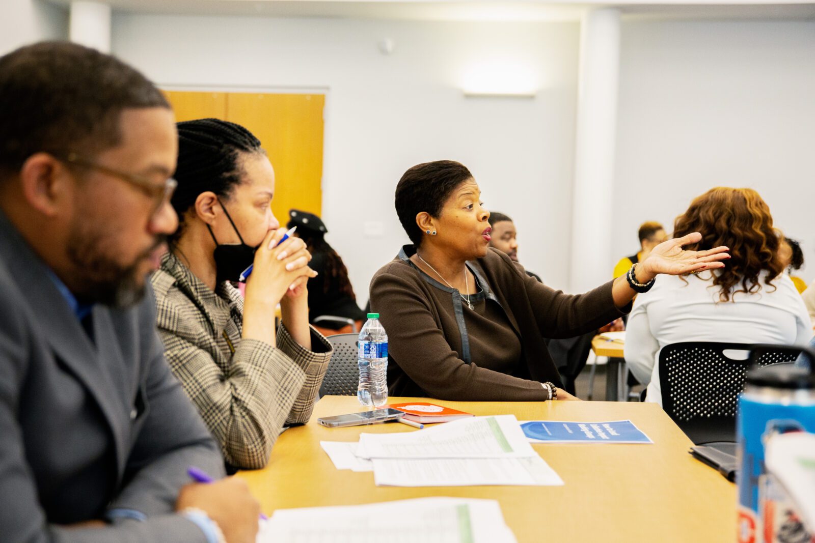 A woman sits at the head of a table, speaking with her arm outstretched at StriveTogether course. Two others sit next to her, looking forward.