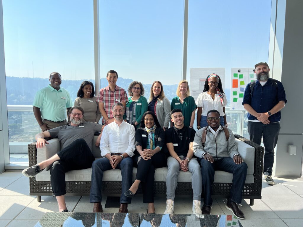 The All Hands Raised board of directors post for a group picture, with a wall of glass windows behind them. Five people sit on a couch and eight people stand behind them.