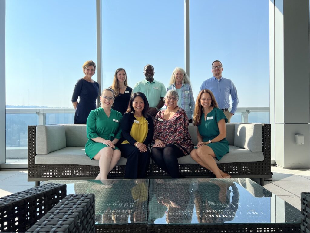 The All Hands Raised staff poses for a group picture, with a wall of glass windows behind them. Four people sit on a couch and five people stand behind them.