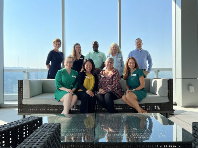 The All Hands Raised staff poses for a group picture, with a wall of glass windows behind them. Four people sit on a couch and five people stand behind them.