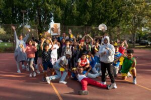 A large group of students pose for a picture. They're standing on an outdoor basketball court, smiling at the picture, some with their hands up and others making peace signs at the camera.