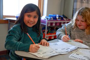 A young student smiles at the camera, holding a pencil to a worksheet