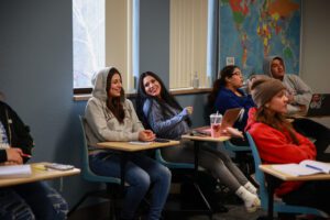 Two high school students sit at desks in the back of a classroom, laughing with each other.