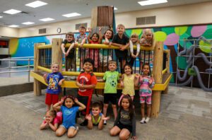 A group of 16 young students pose for a group photo, standing on an indoor playground.