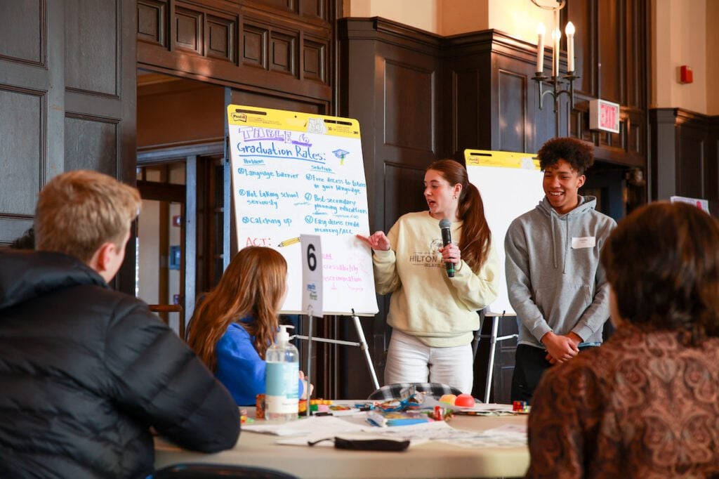 Two students present in front of their peers, reading off of a paper that says 