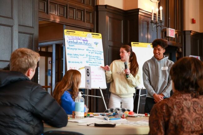 Two students present in front of their peers, reading off of a paper that says "Table 6: Graduation Rates" at the top