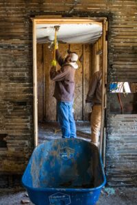 A volunteer works to repair a home damaged by the flood. He's wearing a mask and using a crowbar to pull drywall off the ceiling.