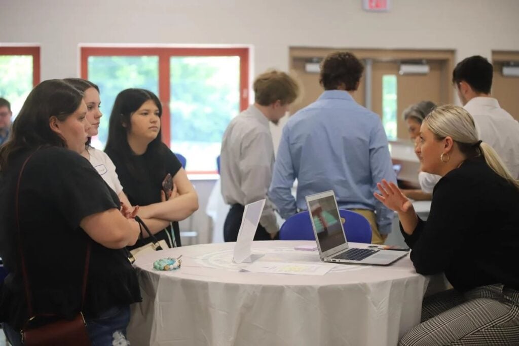 Three high school students stand at an information desk listening to a presenter