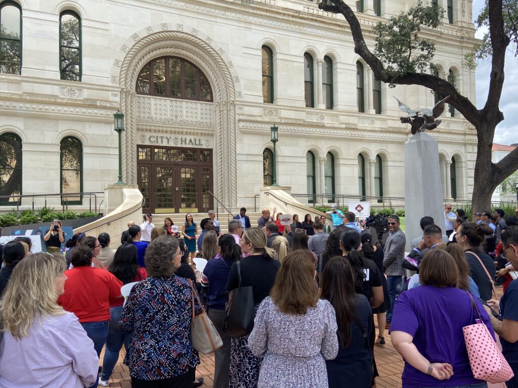 A crowd of people stand facing City Hall