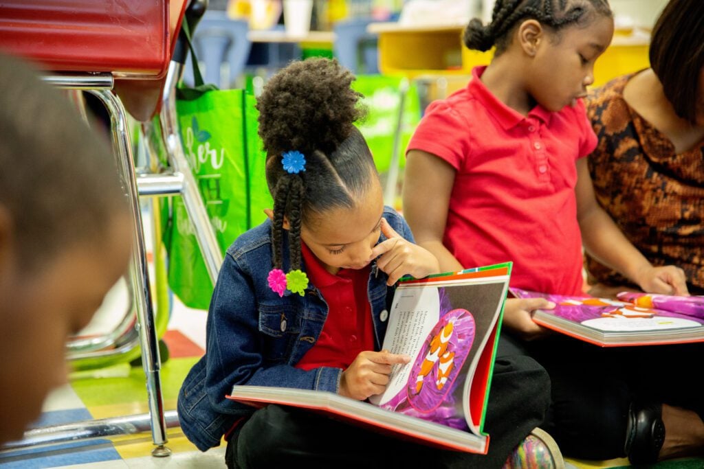 A young girl sits on the ground, reading a book she's holding in her lap.