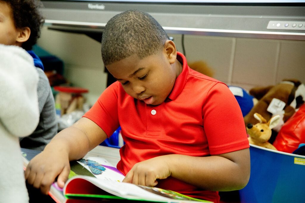 A young boy is reading a book, holding his finger to the page to follow along as he reads.