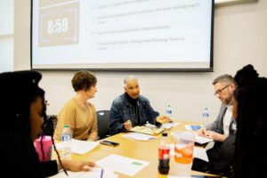 Five adults sit at a table, turned to listen to one woman who is speaking.