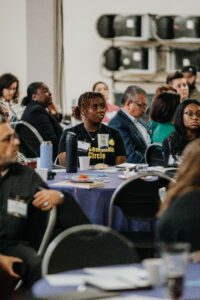 A small crowd sits at tables listening to a presentation. The photo focuses on a young woman at center of the photo.