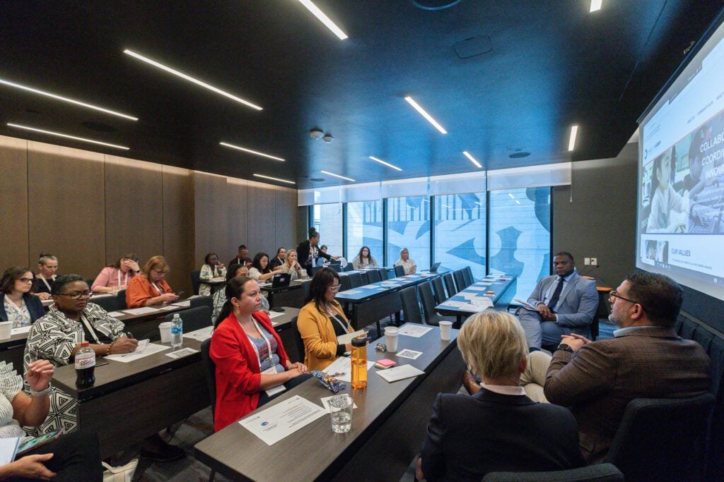 A group of adults sit in rows looking toward three presenters in the front of the room.