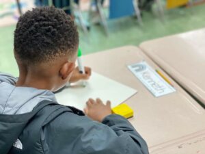 A young student sits at a desk, writing on a piece of paper with a marker.