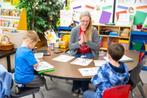 A teacher sits with two young students at a table, working on literacy skills