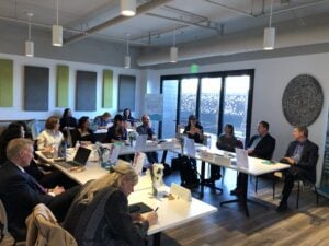 15 adults sit in a room, most sitting at a "U" shaped table. Everyone is turned to face a woman who is speaking.