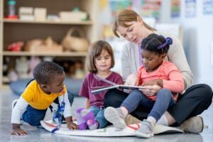 A daycare teacher sits on the floor of the classroom with three young children as she reads a story allowed to them. The children are huddled n closely and are listening attentively.