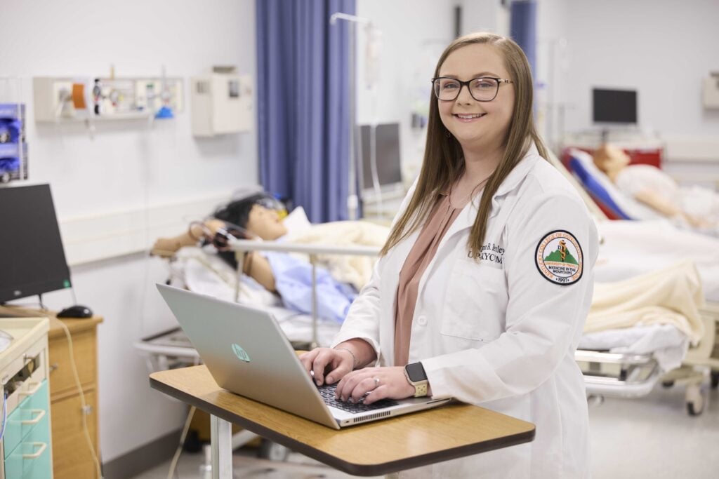 Young woman in a white coat is standing at a desk while smiling at the camera. She is standing in front of hospital beds that are positioned behind her.