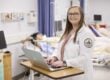 Young woman in a white coat is standing at a desk while smiling at the camera. She is standing in front of hospital beds that are positioned behind her.